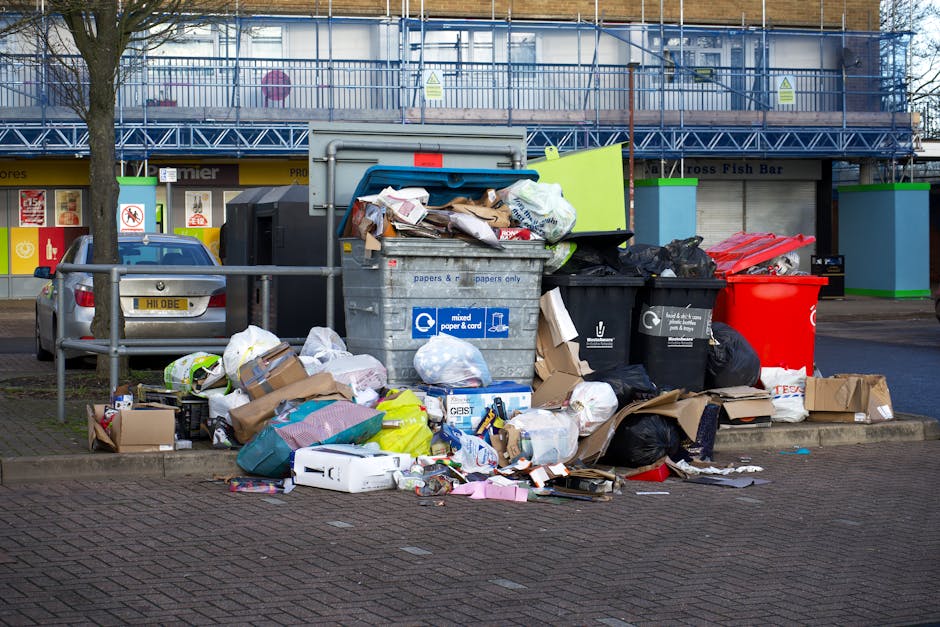 A large collection of mixed waste materials and rubbish bags stacked on a paved street corner in front of a building with blue scaffolding. The waste includes black and black-black plastic bin bags, flattened cardboard boxes, paper and packaging materials, and various discarded items. There is a grey metal recycling bin labeled for mixed paper and card, partially open with some paper waste visible inside. To the left, a silver car is parked behind a metal railing, and a green waste collection container with a small red and white sign is situated nearby. The background features a commercial storefront with a yellow and black sign, along with bright green fire safety cabinets attached to the building facade. The enviroment appears to be an urban area, illustrating improper waste disposal that might be addressed through alternative rubbish removal services offered by companies like House Clearance Brent.