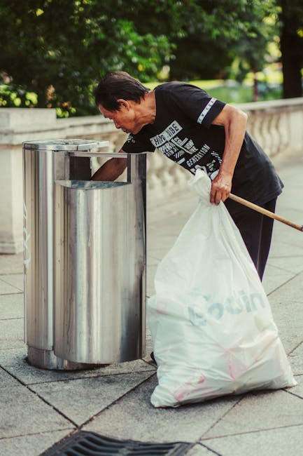 Two large black plastic garbage bags filled with waste are stacked against a dark wooden fence on a paved sidewalk area. One bag appears to be slightly torn, revealing crumpled paper or light debris inside. The bags are situated on the edge of a street with a curb visible in the foreground, and a tree with dense foliage partially visible at the top left corner. The environment suggests an outdoor setting, possibly in a residential area, where private waste collection or rubbish removal services like House Clearance Brent might be involved in managing waste disposal outside of standard municipal refuse systems.
