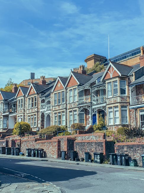 A row of Victorian-style terraced houses with ornate white metal balconies and bay windows, constructed with red brick and grey slate roofing, situated along a street with a brick wall and a line of black wheelie bins in the foreground. The buildings are set on a gentle incline, with some greenery and small trees visible in the front gardens. The sky above is clear with a few wispy clouds, indicating a bright and sunny day. This scene exemplifies typical residential architecture in an urban area, where private property owners or property managers may arrange for independent waste collection services such as those offered by House Clearance Brent, especially for large-scale waste removal or house clearance projects, rather than relying on local authority collections.