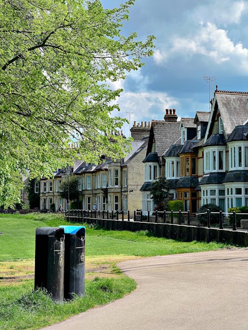 The image shows a residential street with a row of Victorian-style terraced houses featuring bay windows and slate roofs, situated behind a small black metal fence. In the foreground, there is a grassy area with two black rubbish bins positioned near a paved pathway. To the left, a large tree with bright green, fresh leaves extends its branches over the scene, casting dappled shade. The sky above is partly cloudy with patches of blue and white clouds. The environment suggests a quiet suburban neighbourhood, with the presence of waste disposal bins indicating local waste management practices, potentially involving independent collection services like those offered by House Clearance Brent, which provides rubbish removal and house clearance options. Overall, the scene is peaceful, tidy, and depicts typical UK residential street scenery with elements relevant to private waste disposal or rubbish removal services, aligned with the context of alternative waste handling solutions.