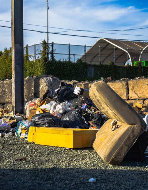 A pile of assorted rubbish and waste materials collected on the side of a gravel surface, including black plastic garbage bags, a yellow plastic container, pieces of broken furniture such as a sofa cushion, and various discarded items. The rubbish is situated near a low stone wall made from large, rough-hewn stones, with some greenery and bushes behind it. In the background, there are utility poles, power lines, and a partially visible modern structure with a curved roof, under a blue sky with some wispy clouds. The scene suggests a location where independent waste collection or private rubbish clearance might be occurring, with the waste temporarily assembled outside for removal or disposal by a service like House Clearance Brent, aligning with the theme of rubbish removal and on-site clearance services.