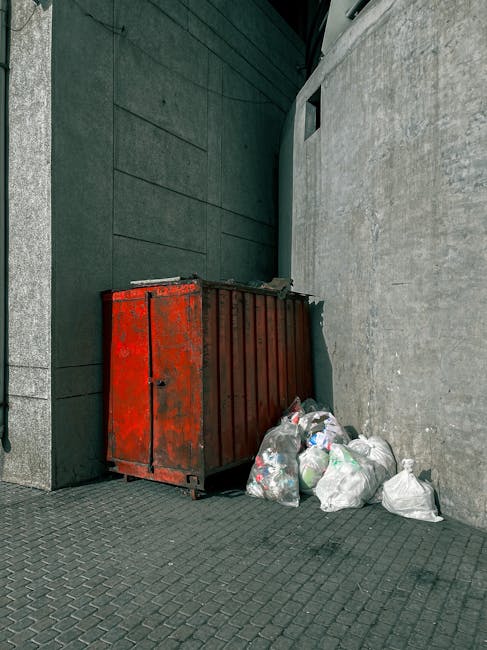 A pile of assorted rubbish and waste materials collected on the side of a gravel surface, including black plastic garbage bags, a yellow plastic container, pieces of broken furniture such as a sofa cushion, and various discarded items. The rubbish is situated near a low stone wall made from large, rough-hewn stones, with some greenery and bushes behind it. In the background, there are utility poles, power lines, and a partially visible modern structure with a curved roof, under a blue sky with some wispy clouds. The scene suggests a location where independent waste collection or private rubbish clearance might be occurring, with the waste temporarily assembled outside for removal or disposal by a service like House Clearance Brent, aligning with the theme of rubbish removal and on-site clearance services.