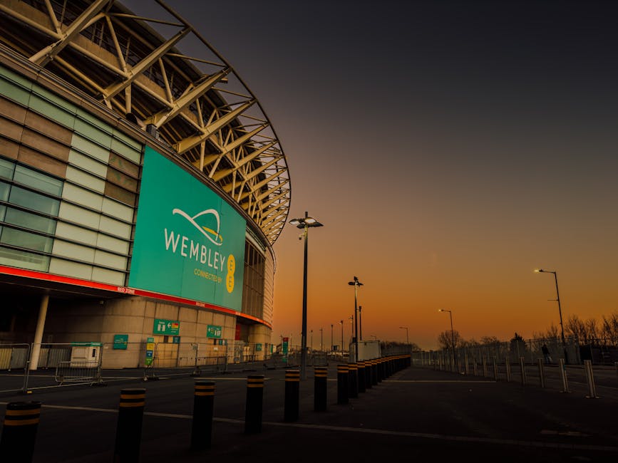 A wide view of Wembley Stadium during a sunset, with the stadium's exterior visible on the left side of the image. The structure features a prominent arched roof with a lattice-like metallic framework and large glass panels, some of which reflect the warm orange and purple hues of the evening sky. A large, turquoise digital billboard displaying the 'WEMBLEY' logo is mounted on the curved façade of the stadium. In the foreground, a paved forecourt area extends outward, lined with black and yellow safety bollards and streetlamps that are turned off. Metal railings and temporary barriers run alongside the pavement, indicating a lack of pedestrian congestion. Several green and white directional signs are positioned near the stadium entrance, and a small truck is parked further down the forecourt. The surrounding environment appears quiet, with distant trees and low structures silhouetted against the colorful sky, emphasizing the stadium's impressive scale and modern architectural design, suitable for supporting private waste handling or on-site clearance activities by companies like House Clearance Brent.