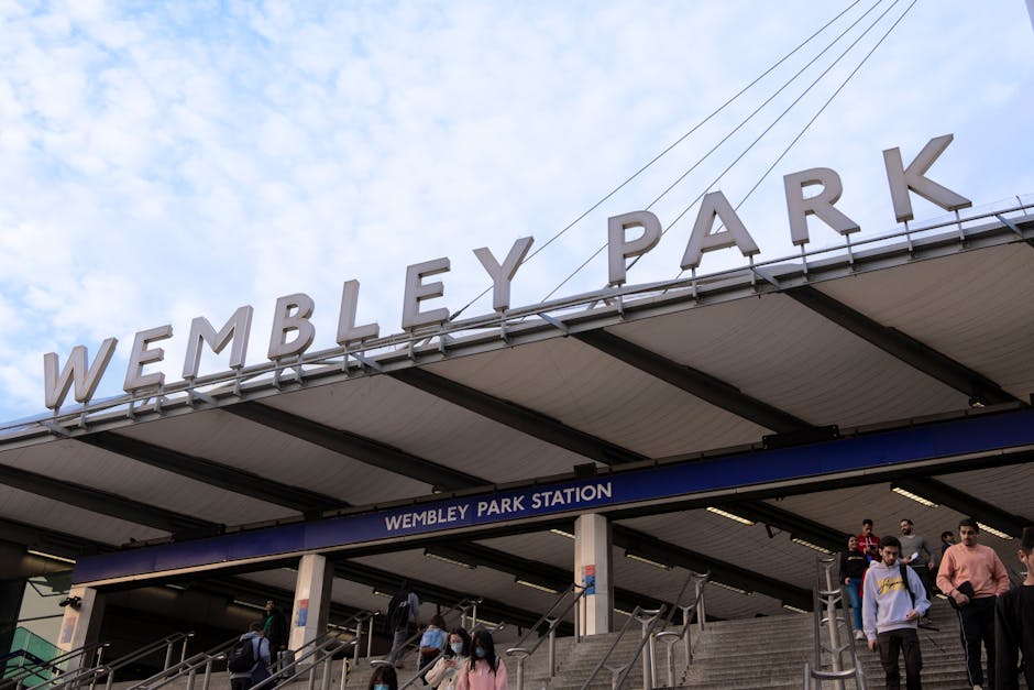 Underneath the large metal signage reading 'Wembley Park' mounted above the entrance to Wembley Park Station, a diverse group of commuters is seen descending concrete stairs. The station's canopy features a white ceiling supported by steel beams, with a deep blue band displaying the text 'Wembley Park Station' beneath the main sign. The sky above is partly cloudy with patches of blue visible. Several individuals are wearing casual clothing, some with backpacks, and a few are wearing face masks. The environment suggests an active transport hub, where passersby are engaged in arriving or departing from the station, illustrating a typical urban scene. This setting aligns with private or independent transportation hubs which often require specific rubbish removal services for station maintenance or upgrades, evidencing a need for efficient waste management solutions in busy transit environments, which companies like House Clearance Brent can support. The composition captures the station’s structure and public interaction, relevant for visual context in articles about waste disposal or station refurbishment projects.