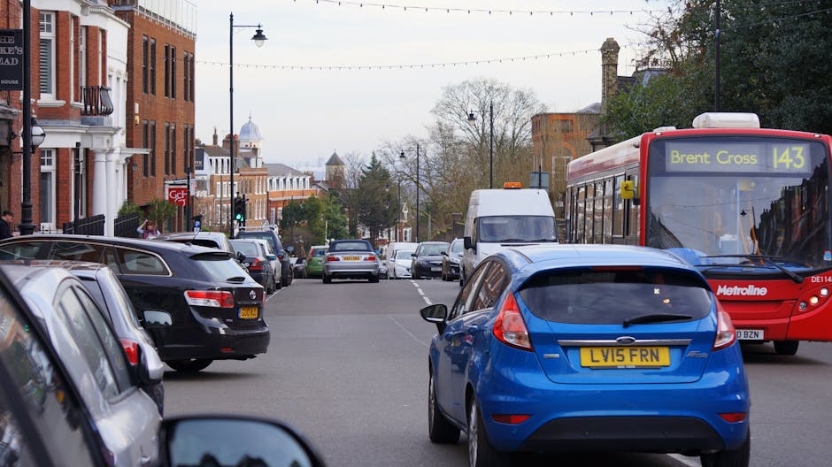 A busy urban street scene in Brent showing parked cars along both sides of the road, including a black hatchback, a blue compact car with a yellow registration plate, and a range of other vehicles. In the background, a red bus with destination signage reading 'Brent Cross 143' is moving along the street, which is lined with brick residential buildings and street lamps. A white van is parked further down the street, surrounded by additional vehicles, with trees and decorative street lighting overhead. The environment appears typical of a city neighbourhood, with a mix of modern and historic architecture, and the scene captures the daily flow of traffic and public transportation. House Clearance Brent may handle waste removal for such areas, including alternative rubbish collection managed outside of local authority services.